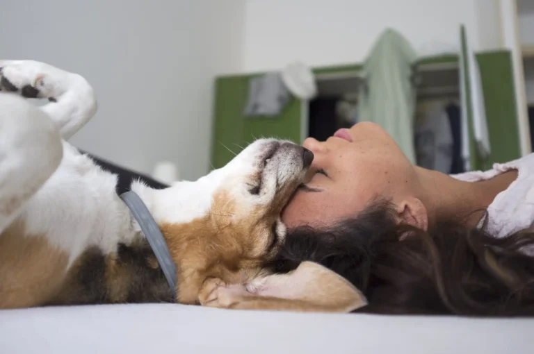 Nell'immagine una ragazza sdraiata su un letto appoggia il suo viso contro il muso di un cagnolino, anche lui sdraiato e